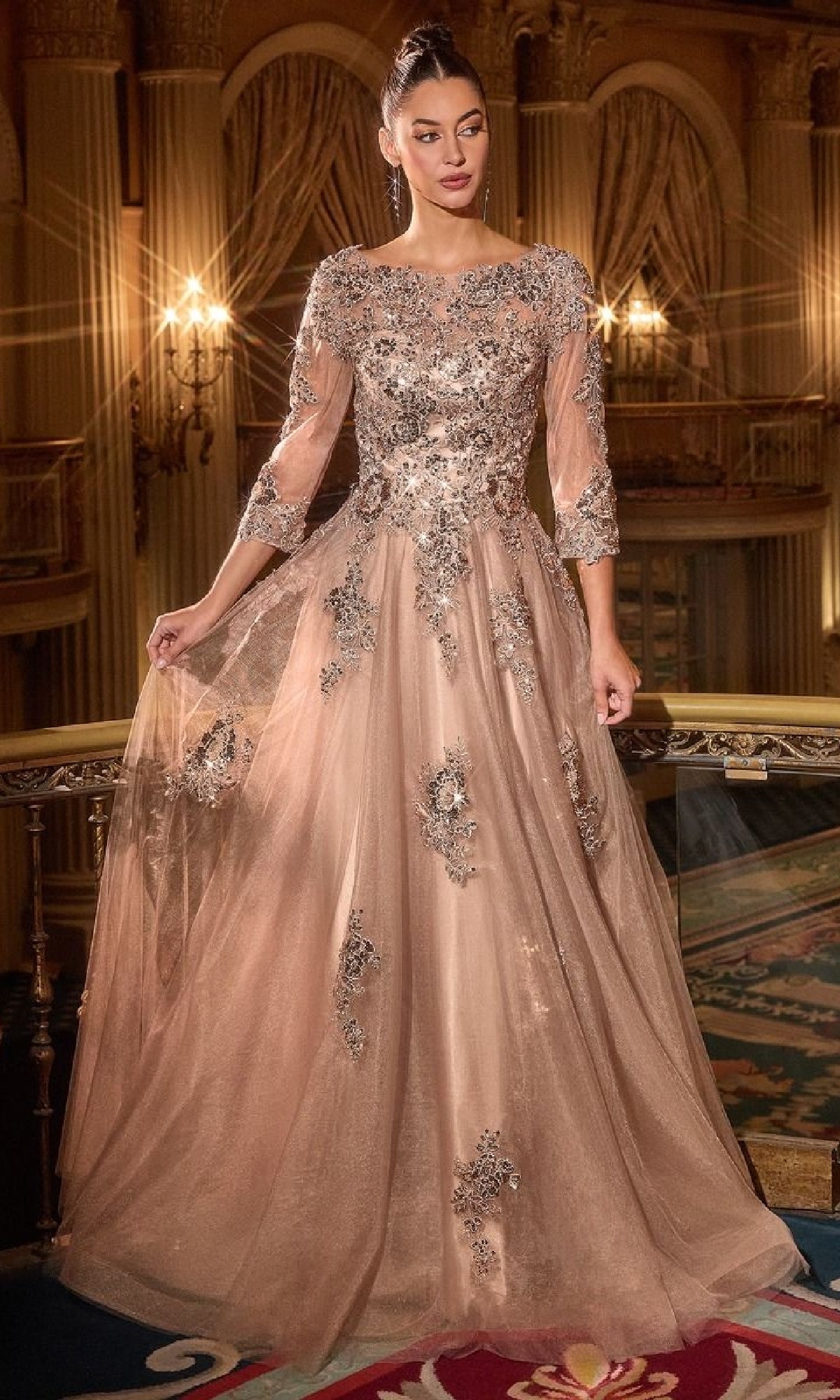 A woman in the Ladivine J883 Long Sleeve Long Formal Dress, featuring a champagne hue with silver floral accents and sheer sleeves, stands in an ornate, elegant interior.